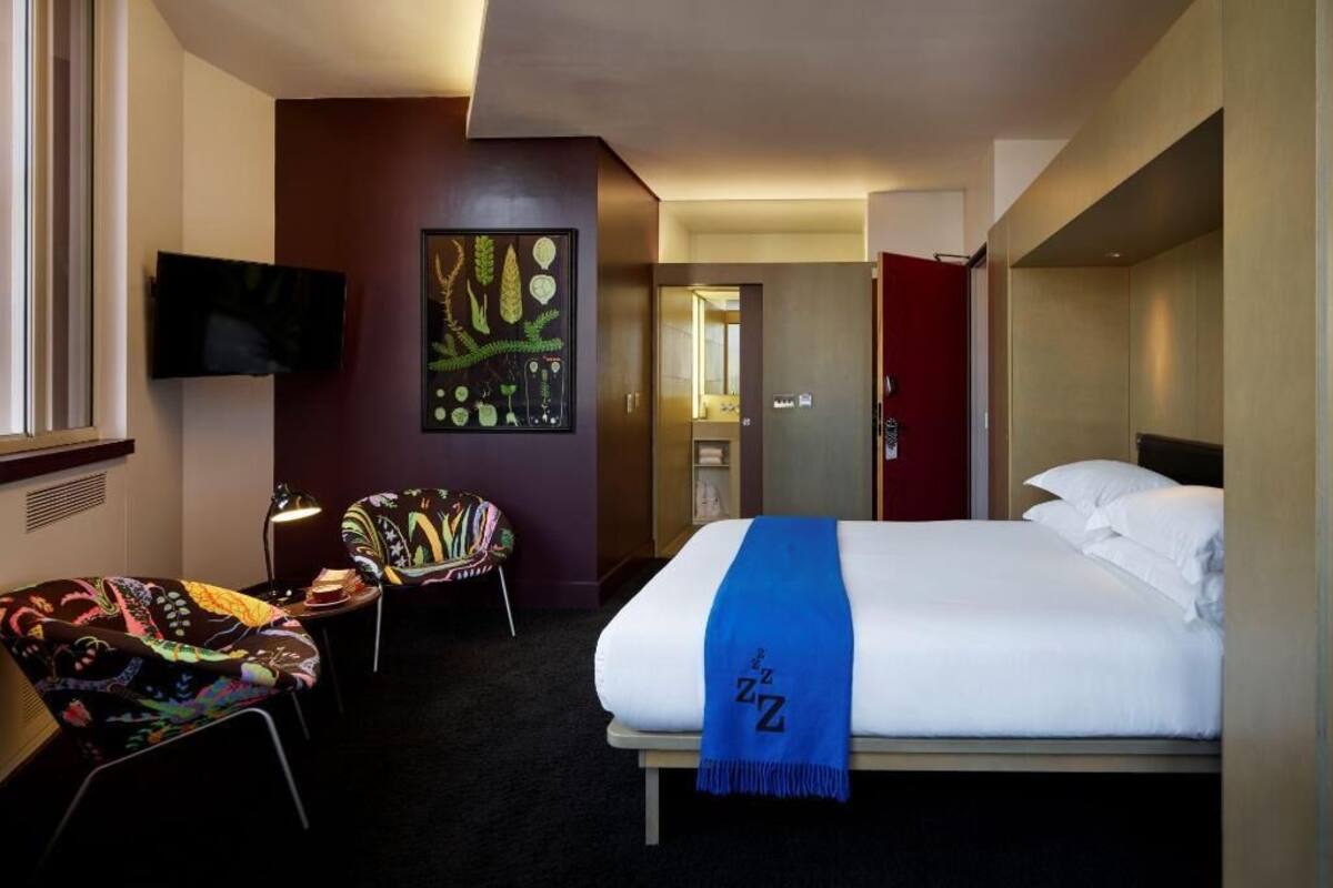 A hotel room with a bed, a TV mounted on the wall, a chair, and a painting.