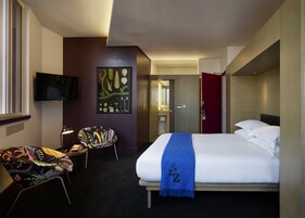 Ein Hotelzimmer mit einem Bett, einem Sessel, einem Schreibtisch, einem Fernseher und einem Wandbild.