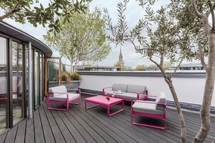 A rooftop terrace with a modern outdoor seating area, including a pink table and grey sofa, overlooking a cityscape with a church spire.