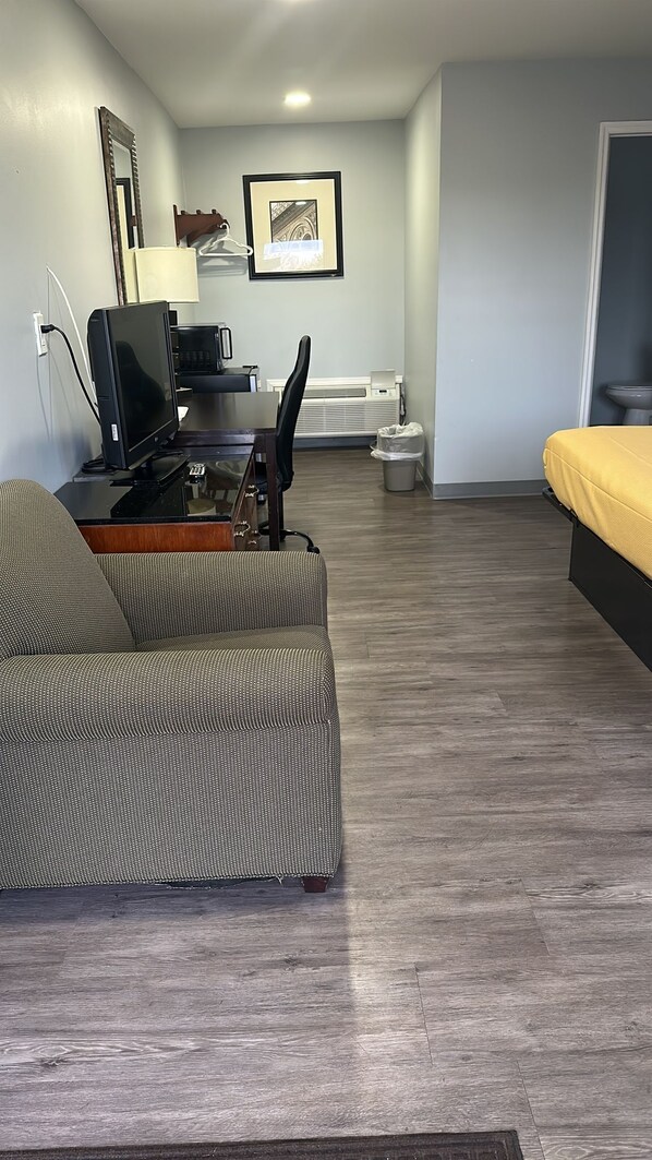A compact living space with a sofa, a small desk with a computer, a chair, a bed, a framed picture on the wall, and a trash bin.