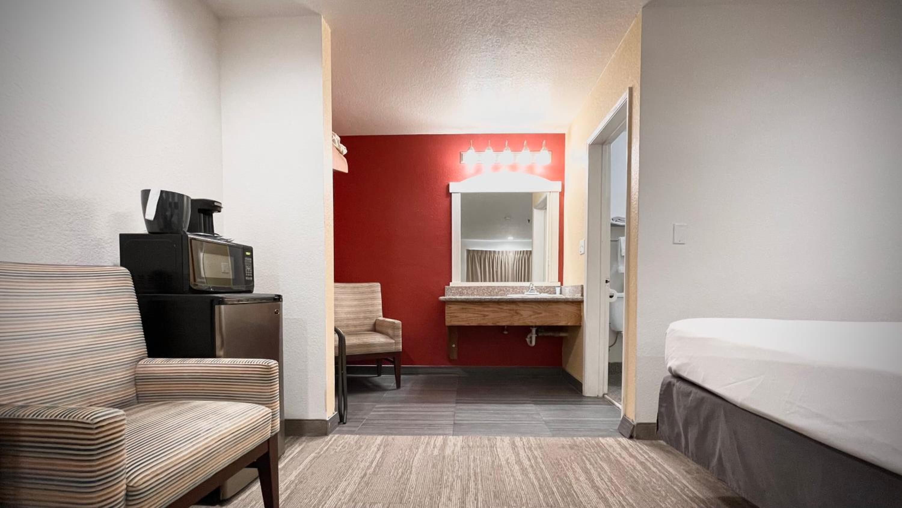 A hotel room with a bed, a small desk with a microwave, a chair, a vanity with a mirror, and a red accent wall.