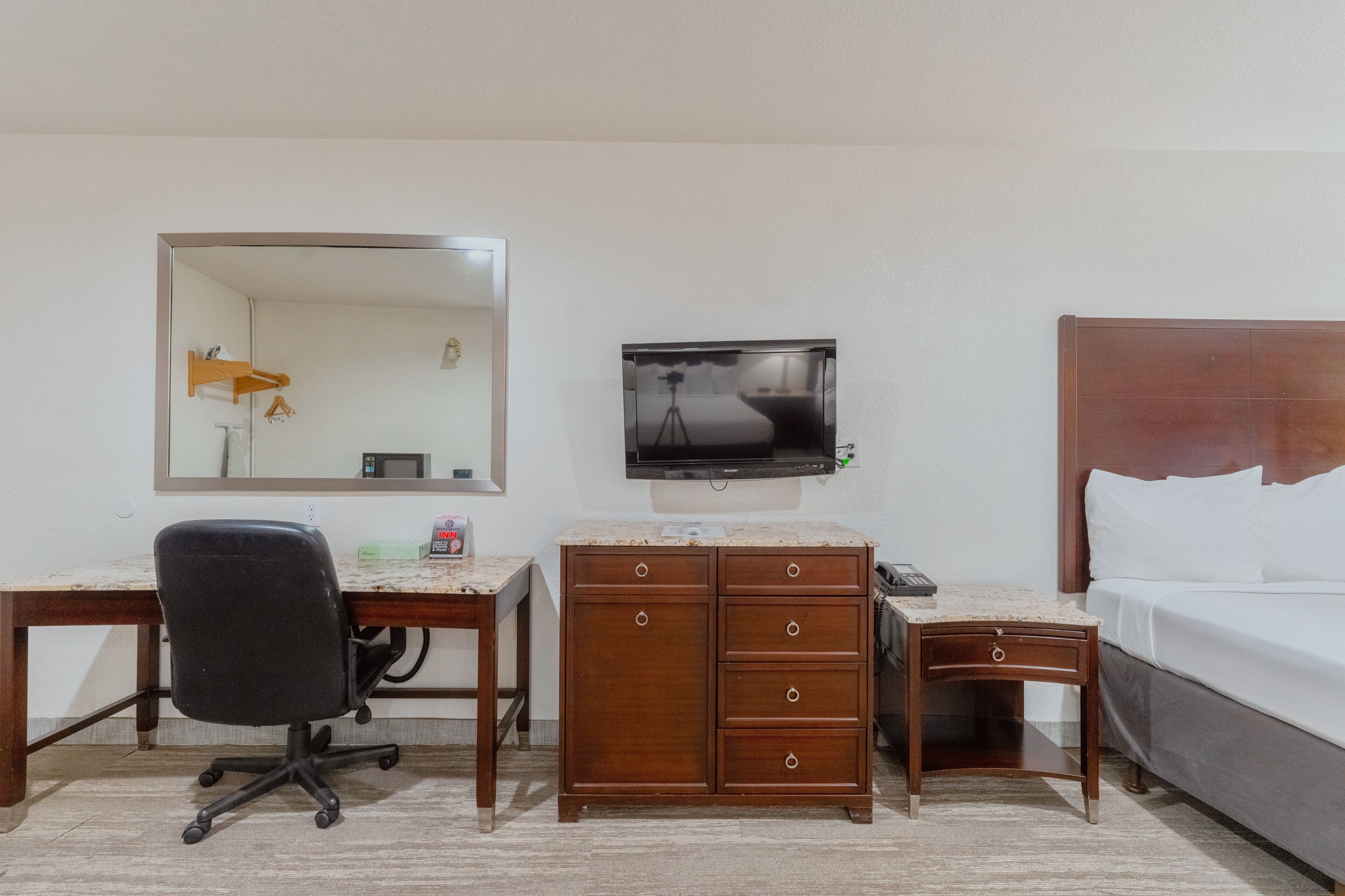 A hotel room with a wooden desk, a black office chair, a TV mounted on the wall, a wooden nightstand, and a bed with white linens.