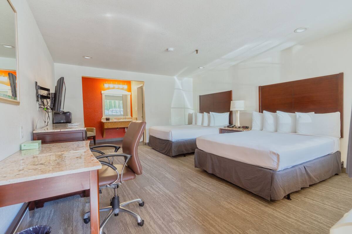 A hotel room with two beds, a desk, a chair, a TV, and a bathroom with a mirror and sink.