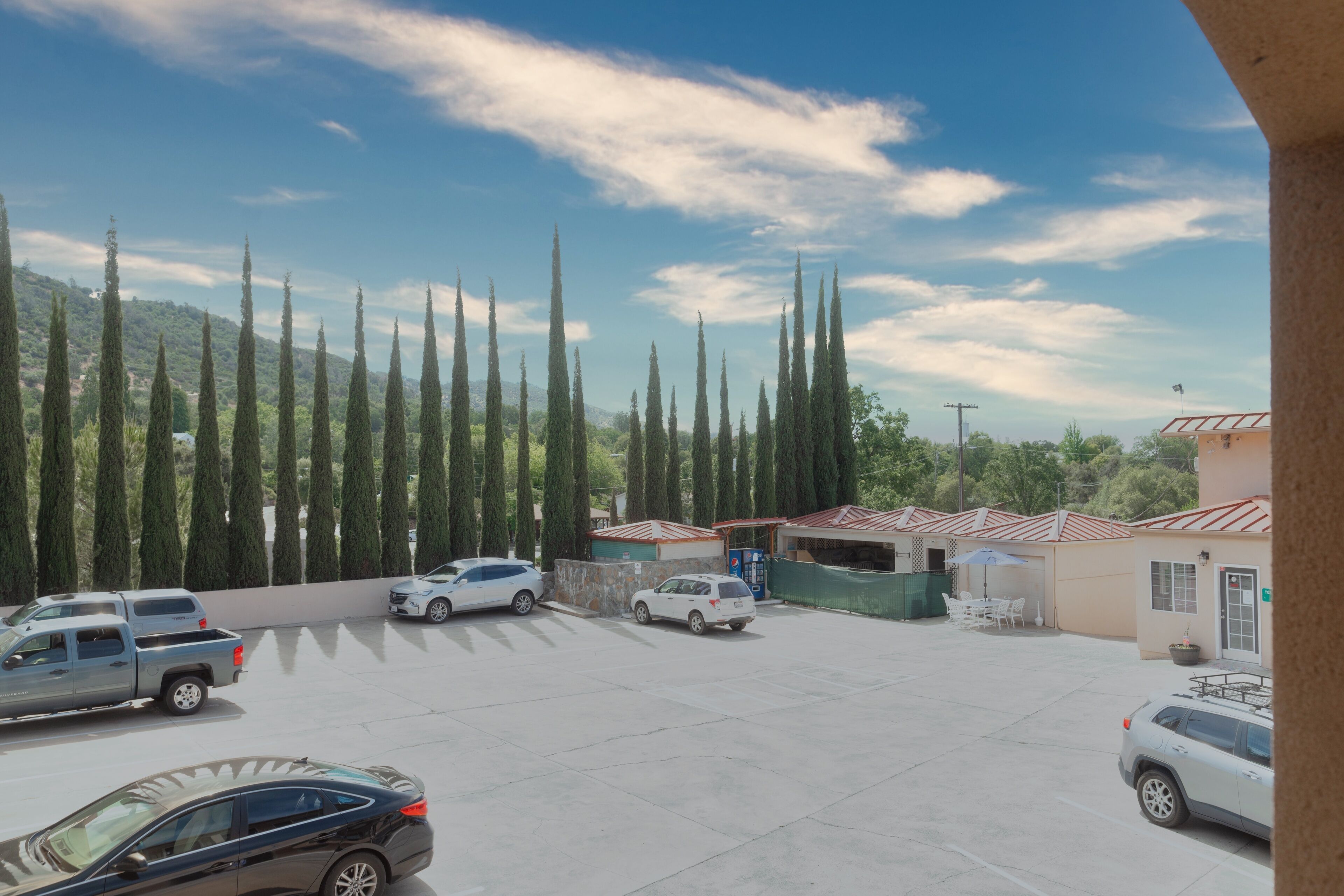 A parking lot with several cars, a building with a red roof, and tall cypress trees in the background.