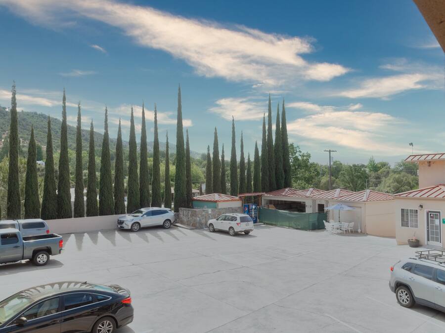 Un aparcamiento con varios coches, un edificio de tejado rojo y altos cipreses al fondo.