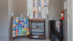 Habitació petita amb finestra, prestatge de fusta amb revistes, nevera negra i extintor.