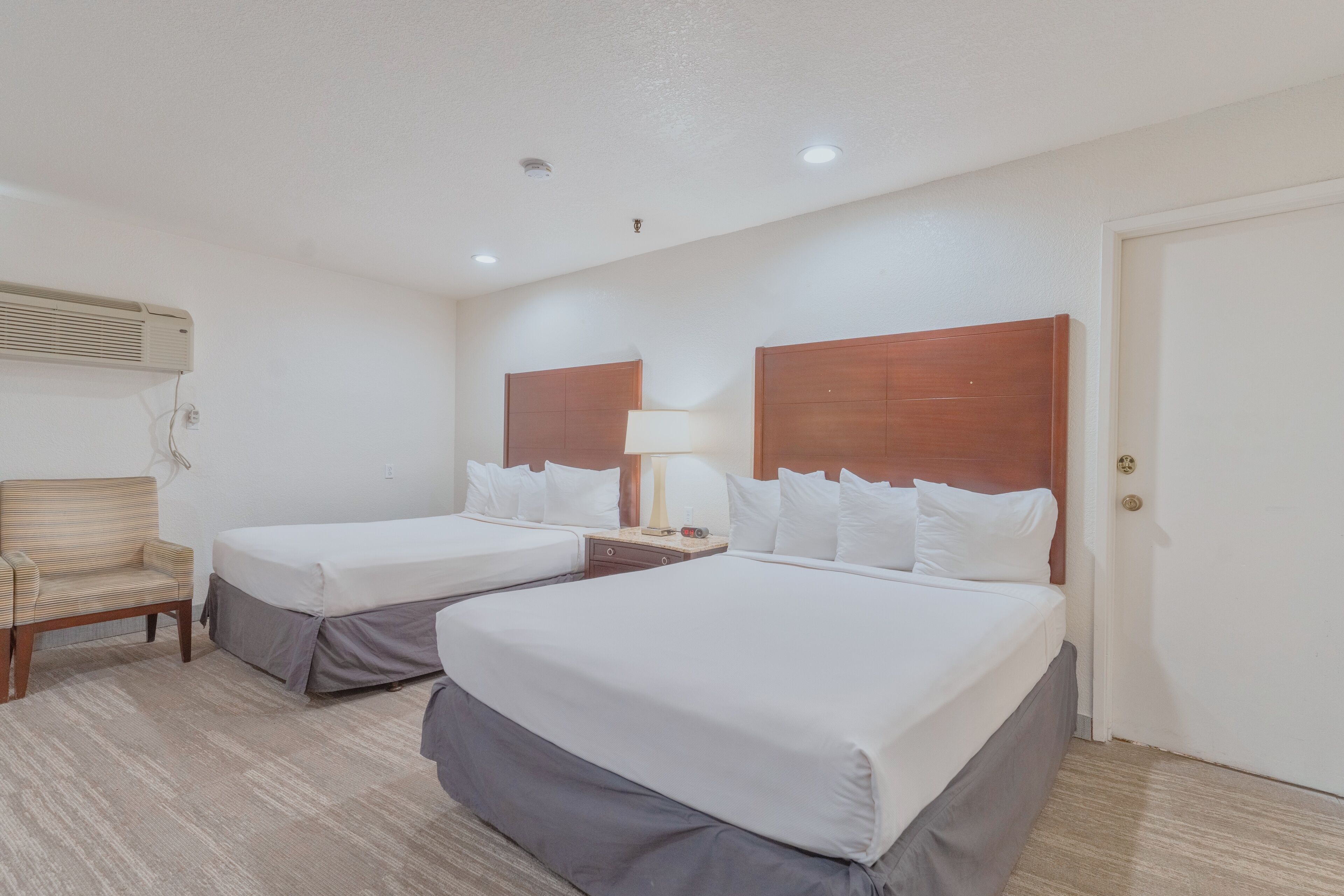 A hotel room with two single beds, a chair, a lamp, and a wall-mounted air conditioner.