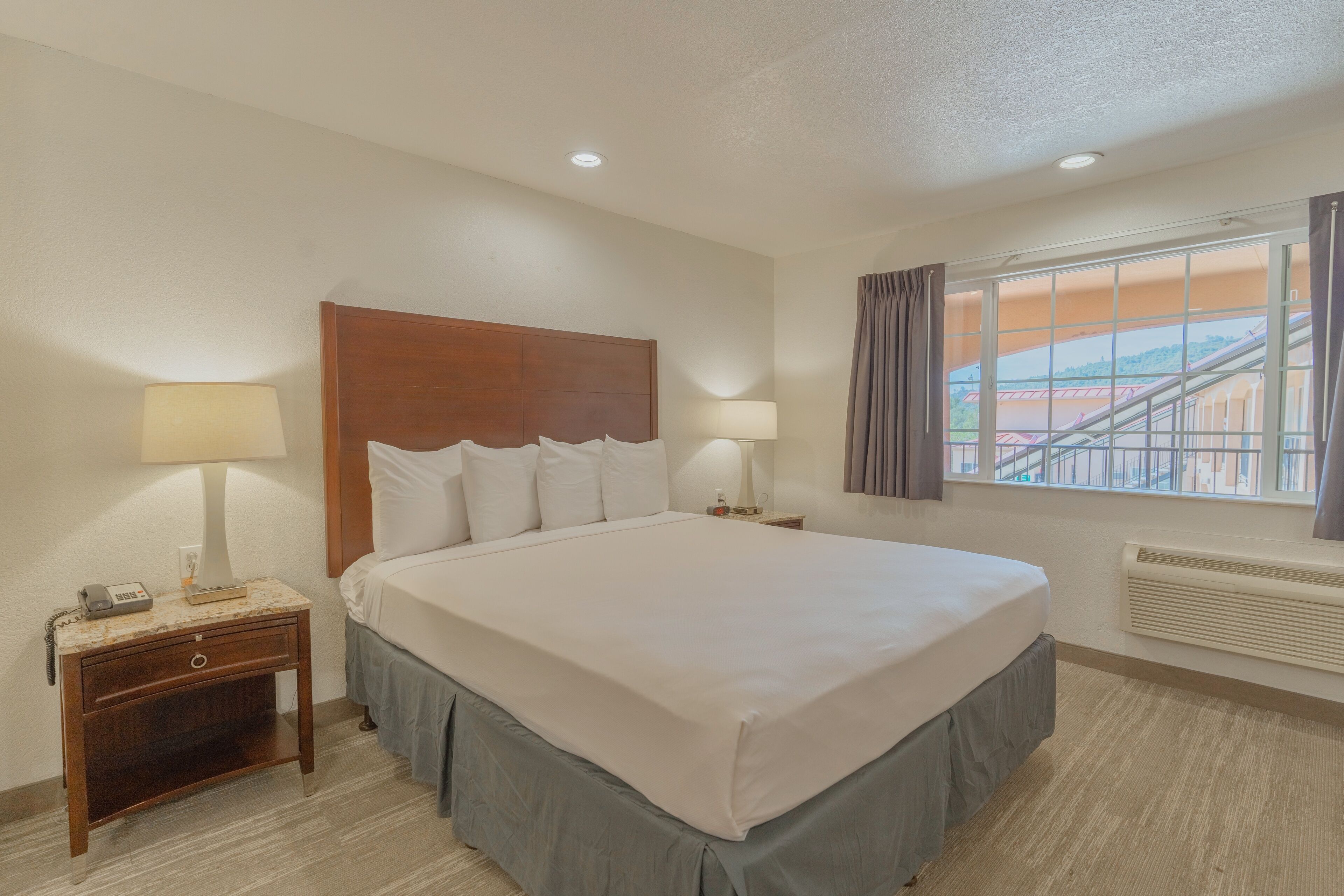 Une chambre d’hôtel avec un grand lit, deux tables de chevet avec des lampes, une table de nuit, une fenêtre avec des rideaux et une vue sur un bâtiment à l’extérieur.