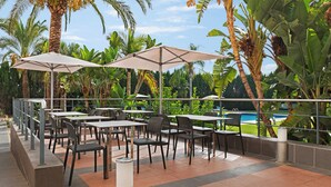 An outdoor patio with tables and chairs, surrounded by palm trees and a pool view. - Ramada by Wyndham Valencia Almussafes (Almussafes)
