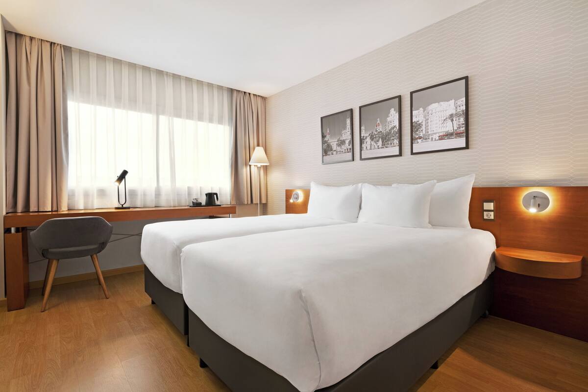 Une chambre d’hôtel avec deux lits, un bureau, une chaise et une grande fenêtre avec des rideaux.