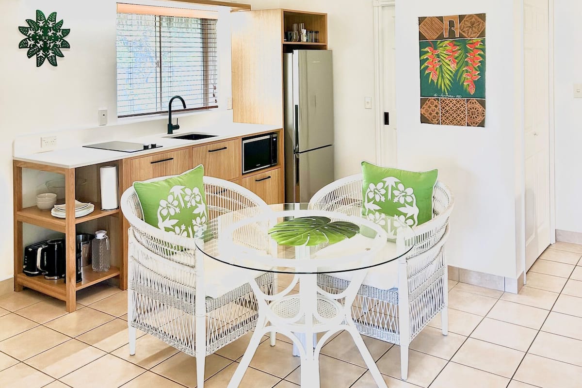 A compact kitchen with a refrigerator, microwave, and wooden cabinets. There is a dining area with a glass table and white wicker chairs, and a wall-mounted light fixture.