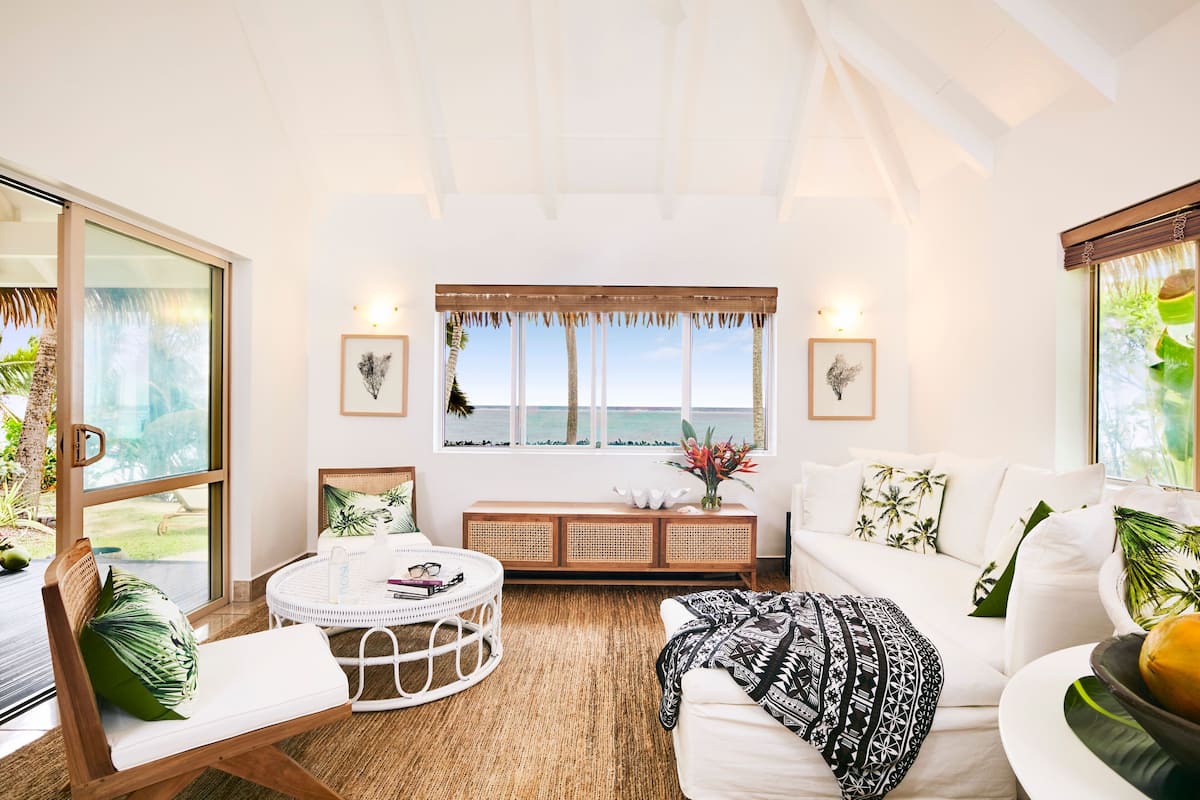 A spacious living room with a large window overlooking the ocean, a white sofa, a round coffee table, and a wooden bench with a plant.