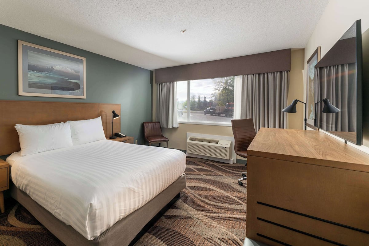 A hotel room with a large bed, a desk, a chair, a nightstand, a lamp, a picture on the wall, and a window with curtains.