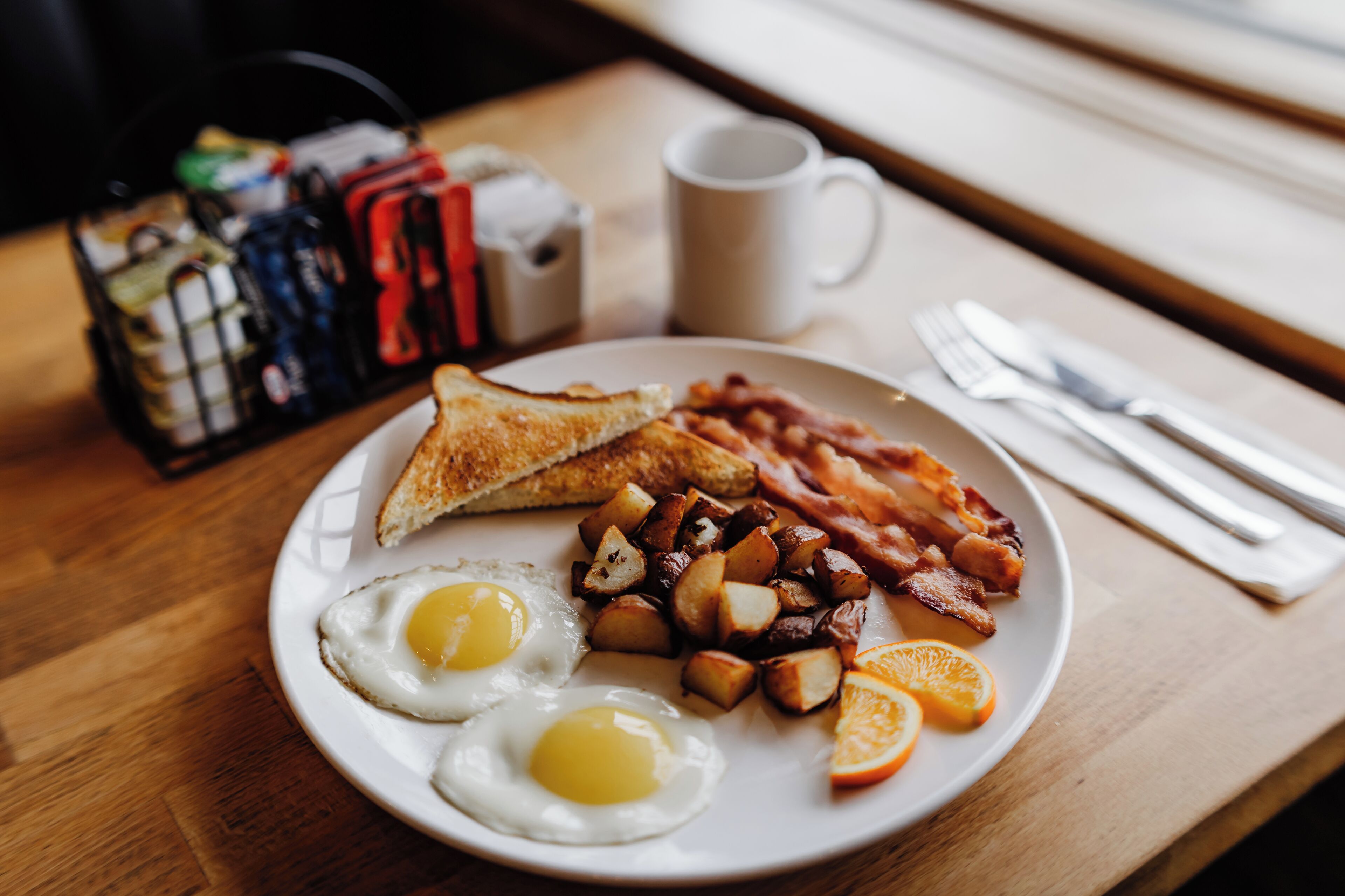 A breakfast plate with eggs, toast, bacon, potatoes, and orange slices on a wooden table.