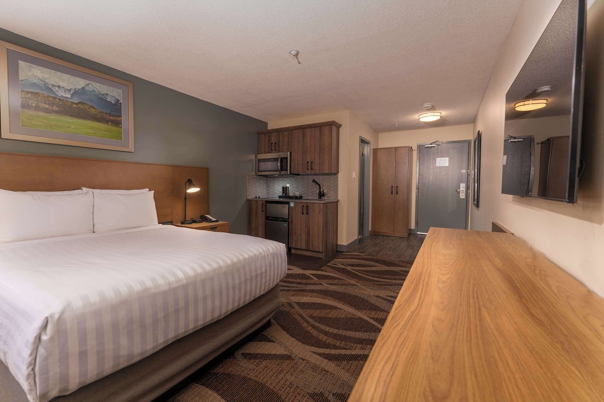 A hotel room with a bed, a wooden desk, a kitchenette, and a bathroom.