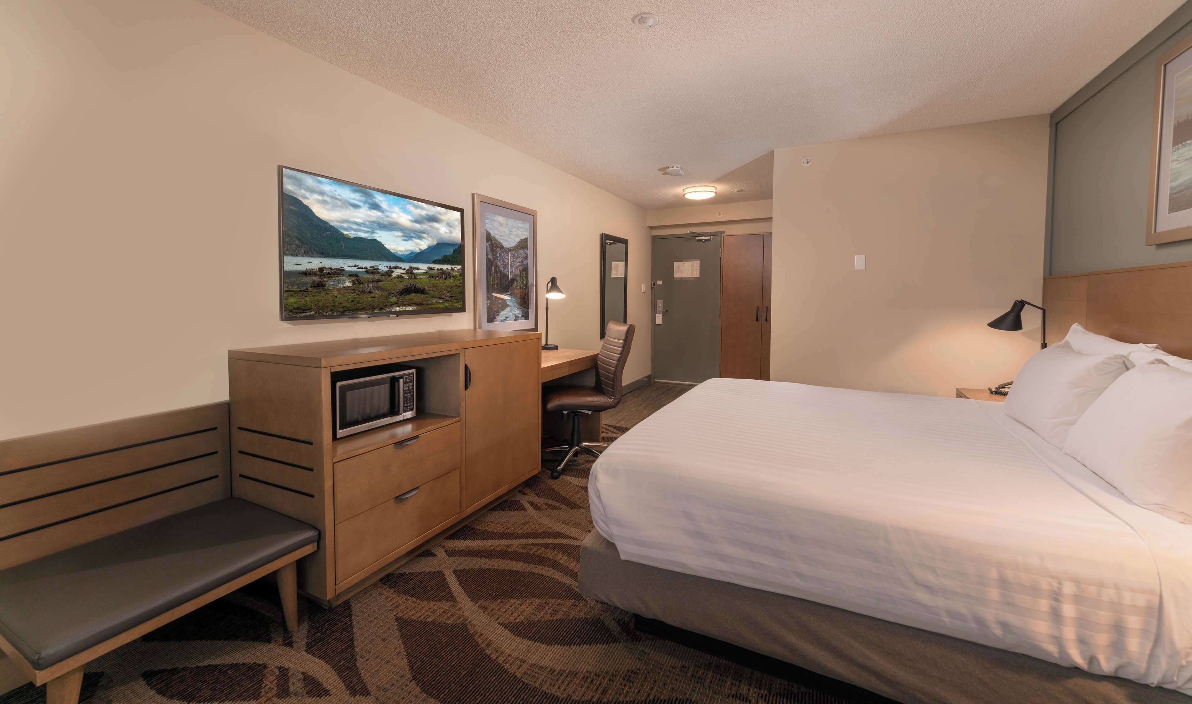A hotel room with a bed, a desk with a computer, a microwave, a bench, and two framed pictures on the wall.