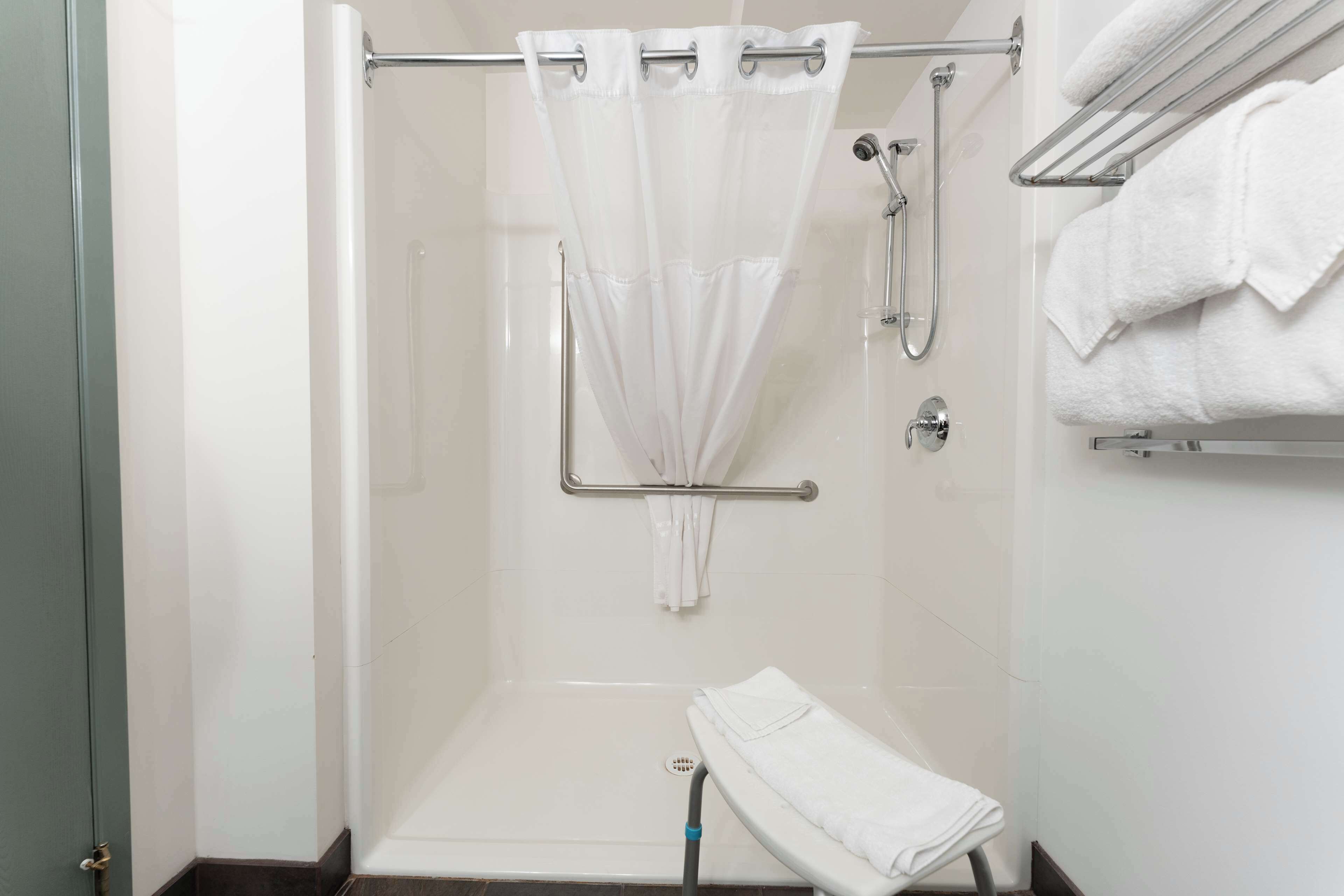 Zona de ducha con puerta de vidrio, cortina de ducha blanca, barra de apoyo y toallero con toallas.