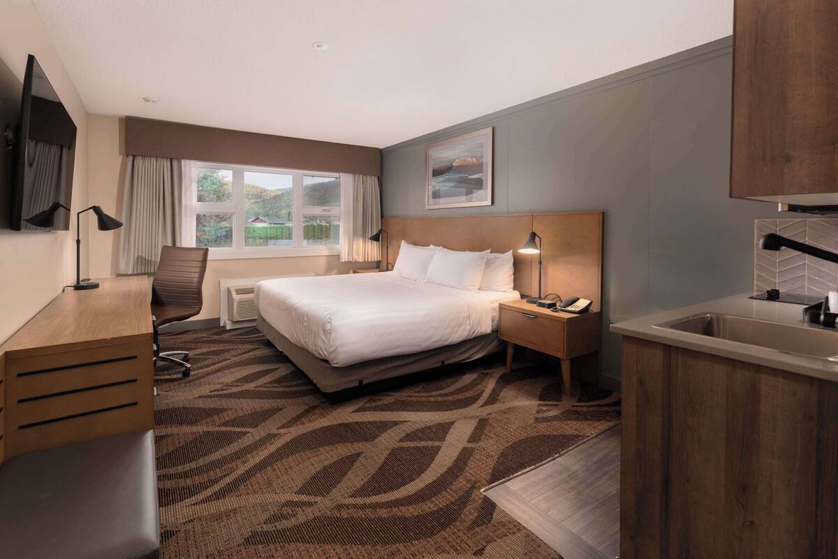 A hotel room with a large bed, a desk, a chair, a nightstand with a phone, a window with curtains, a painting on the wall, and a kitchen area with a sink and countertop.