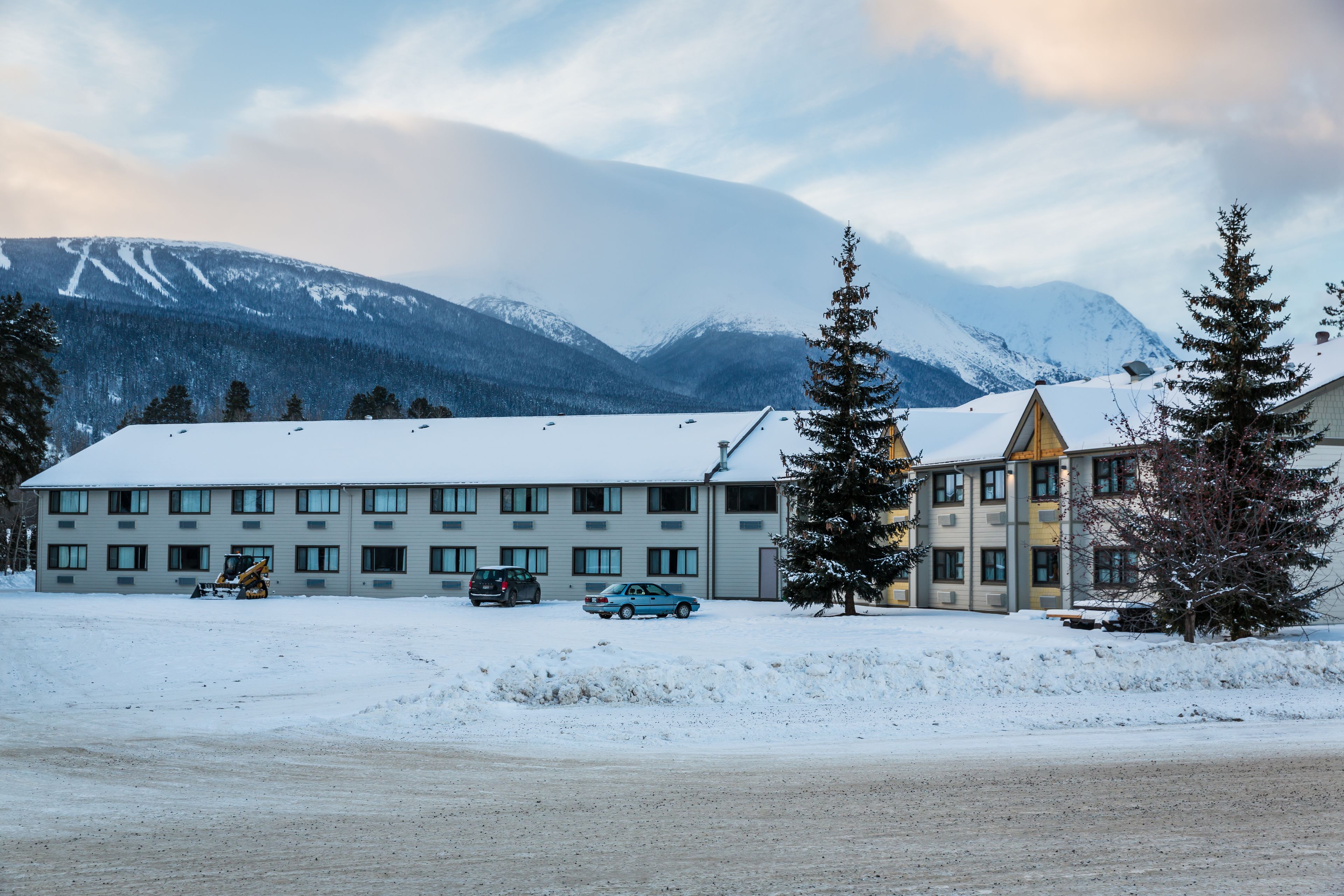 Ein Hotel mit verschneitem Parkplatz, mehreren Fahrzeugen und einem Bergpanorama im Hintergrund.