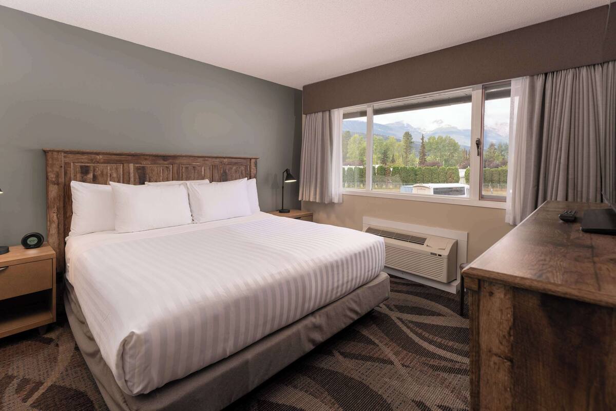 A hotel room with a large bed, a desk, a chair, a lamp, a heater, and a window with curtains.