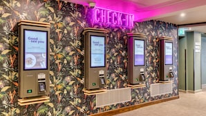 A check-in area with three self-service kiosks against a floral wallpaper backdrop, a neon 'CHECK-IN' sign, and a visible exit door. - Premier Inn London Docklands - Excel (London)