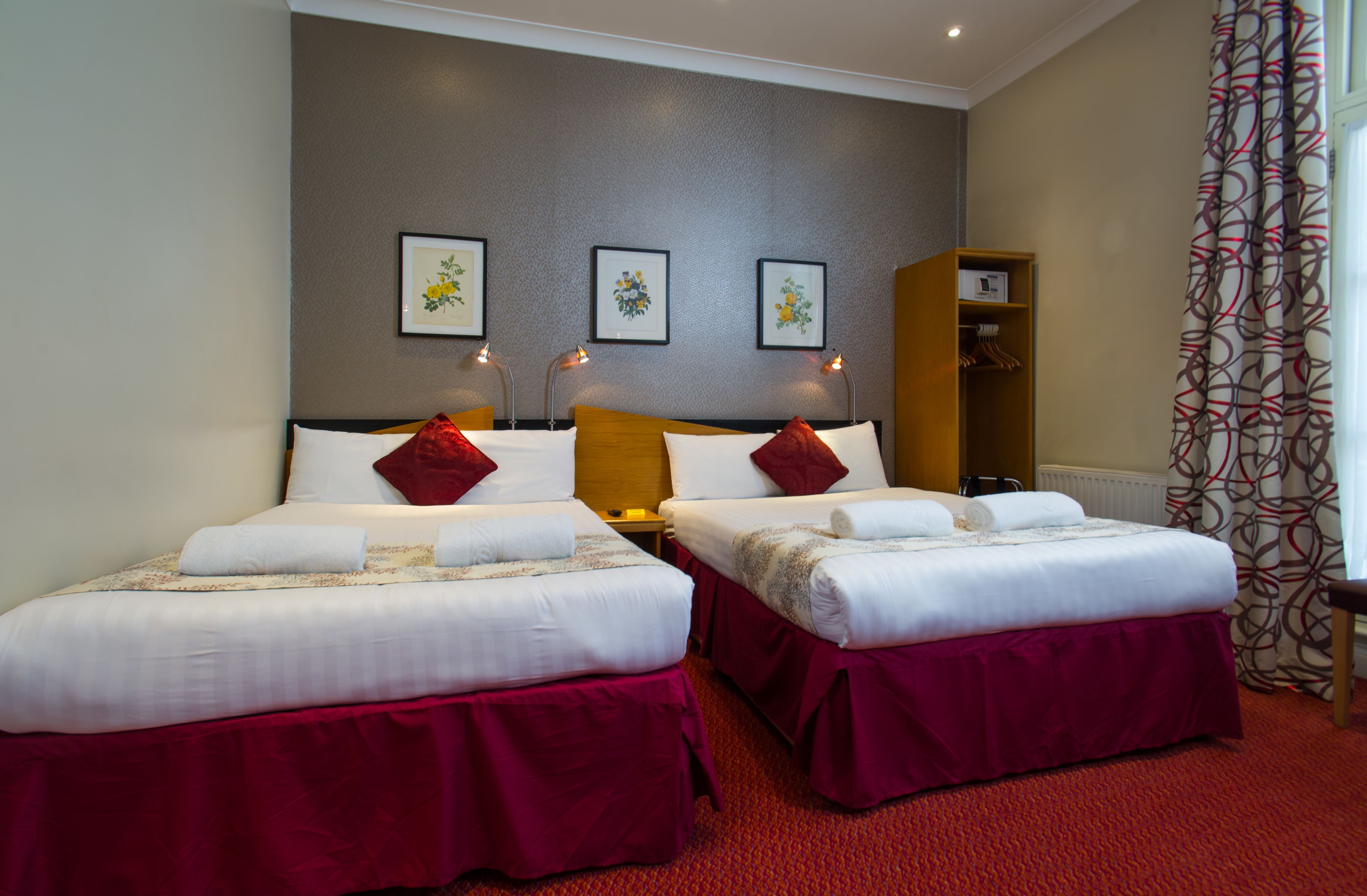 Ett hotellrum med två sängar, röda sängöverkast, en garderob i trä och inramade konstverk på väggen.