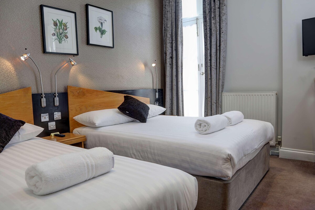 A hotel room with two beds, a wooden headboard, a nightstand, a framed picture, a radiator, and a TV on the wall.