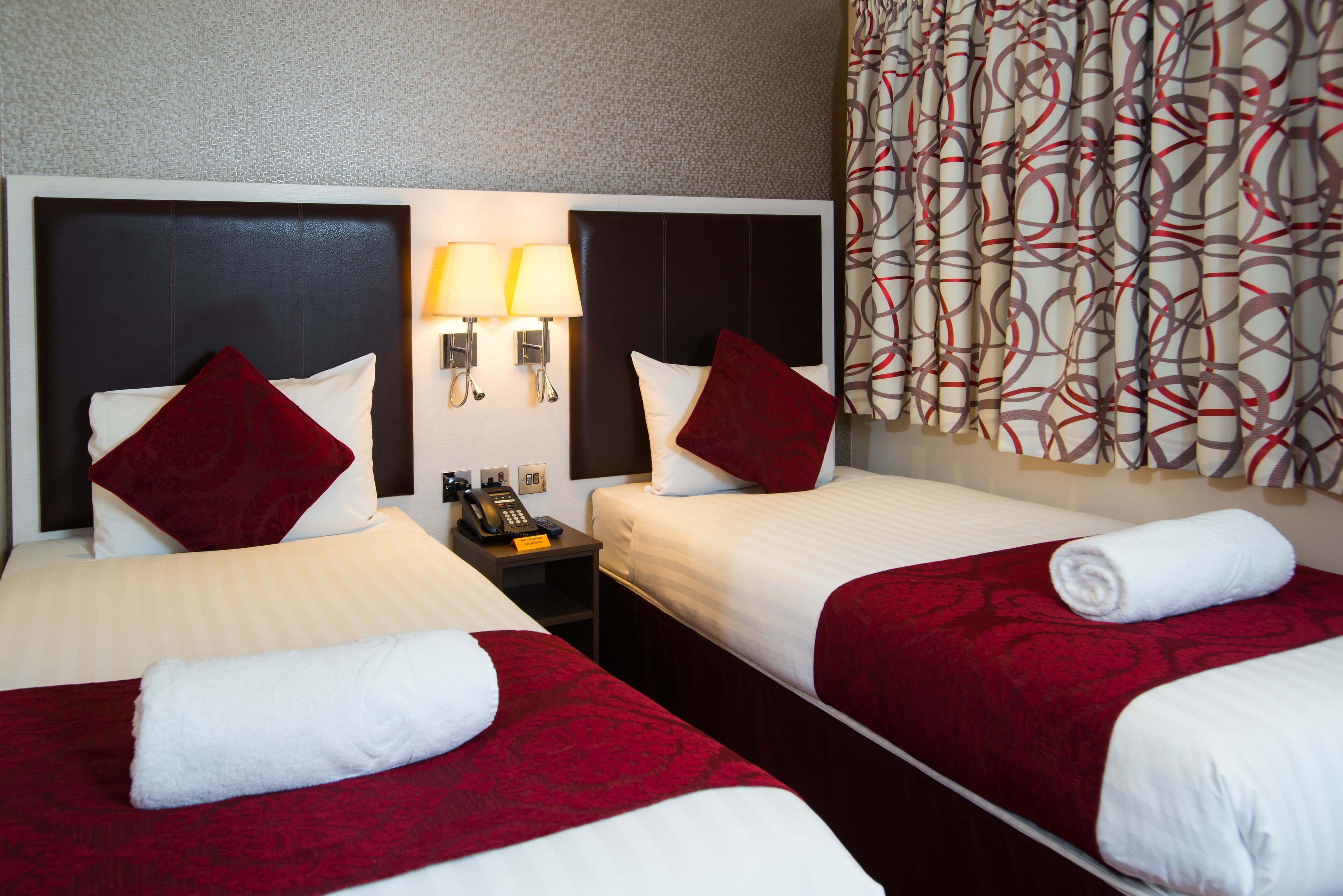A hotel room with two beds, each with a red bedspread and white pillows. There is a nightstand between the beds with a telephone and a lamp. The room has a patterned curtain and a headboard with a light switch.