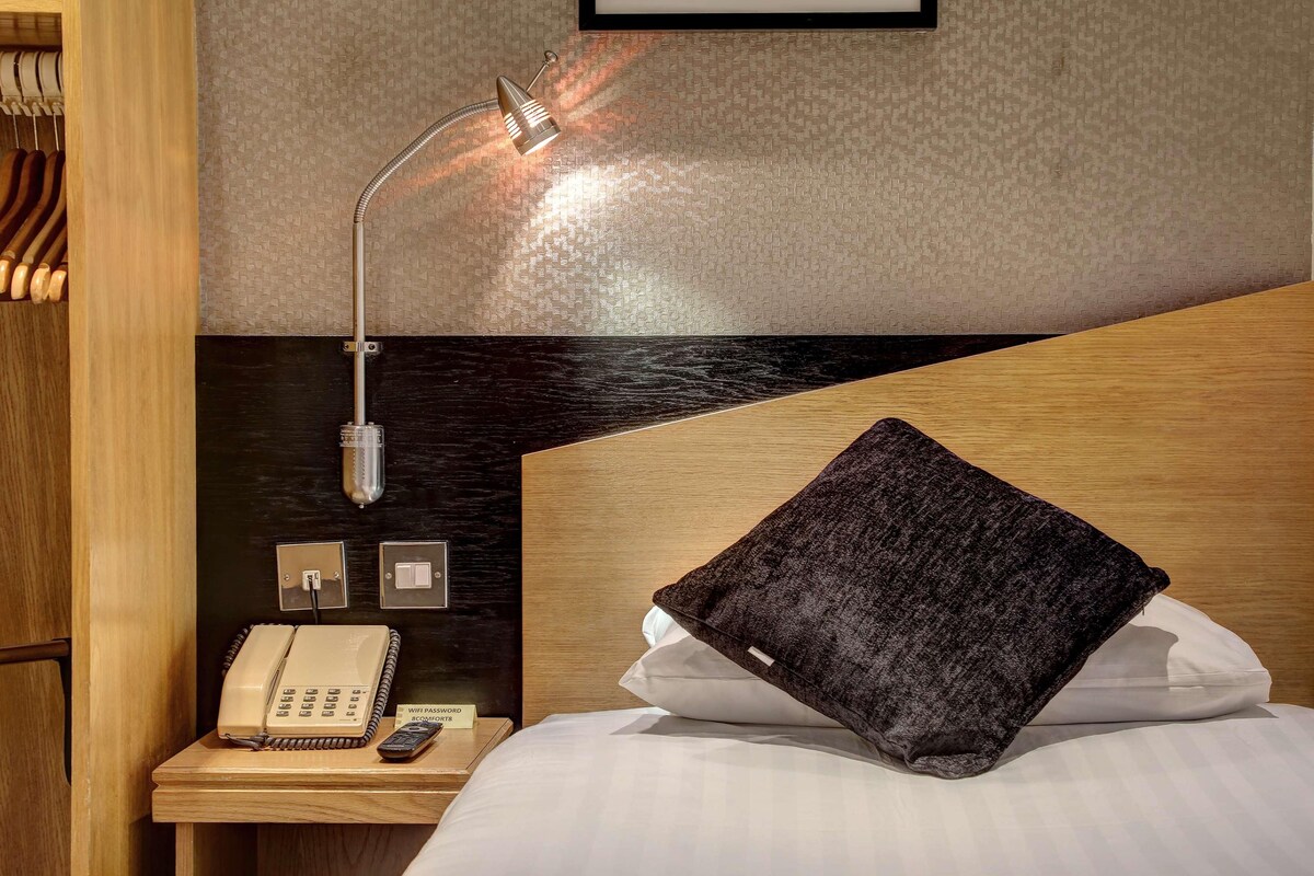 A hotel room with a bed, a bedside table with a telephone, a remote control, and a sign indicating a WiFi password.