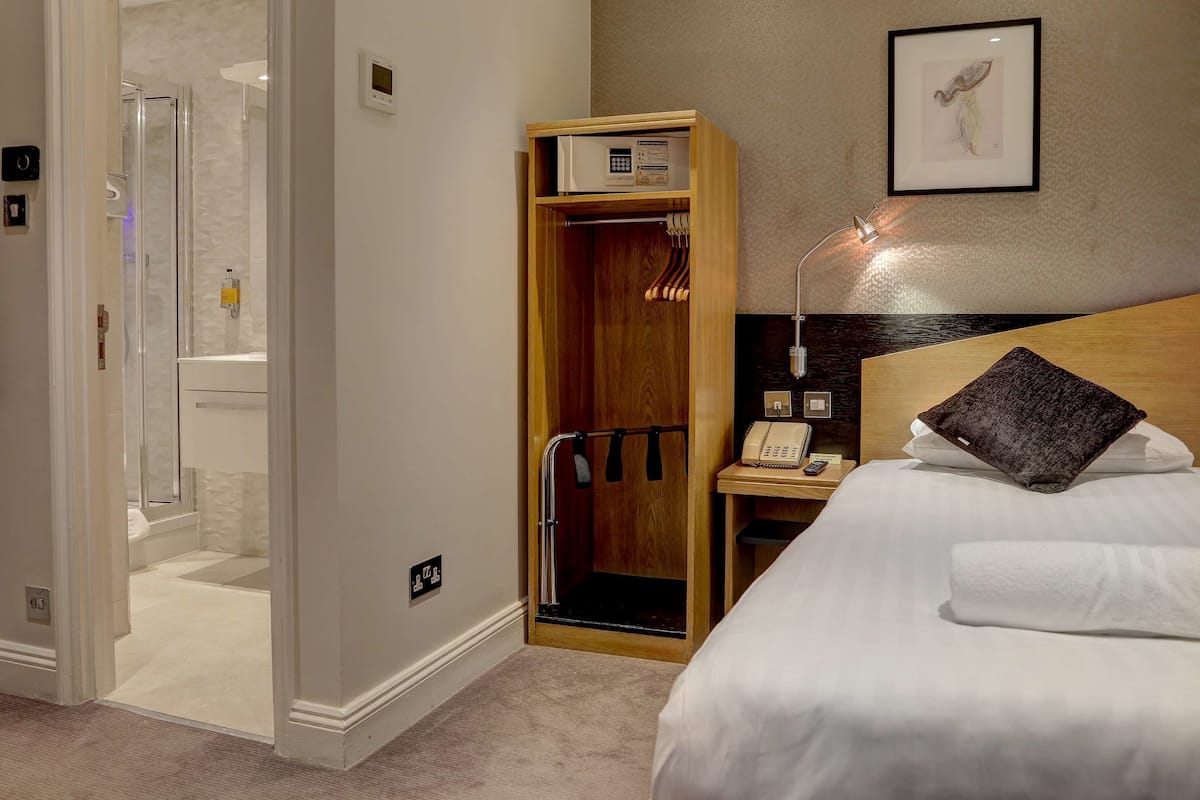 A hotel room with a bed, a wooden wardrobe, a shower area, and a bedside table with a lamp and phone.