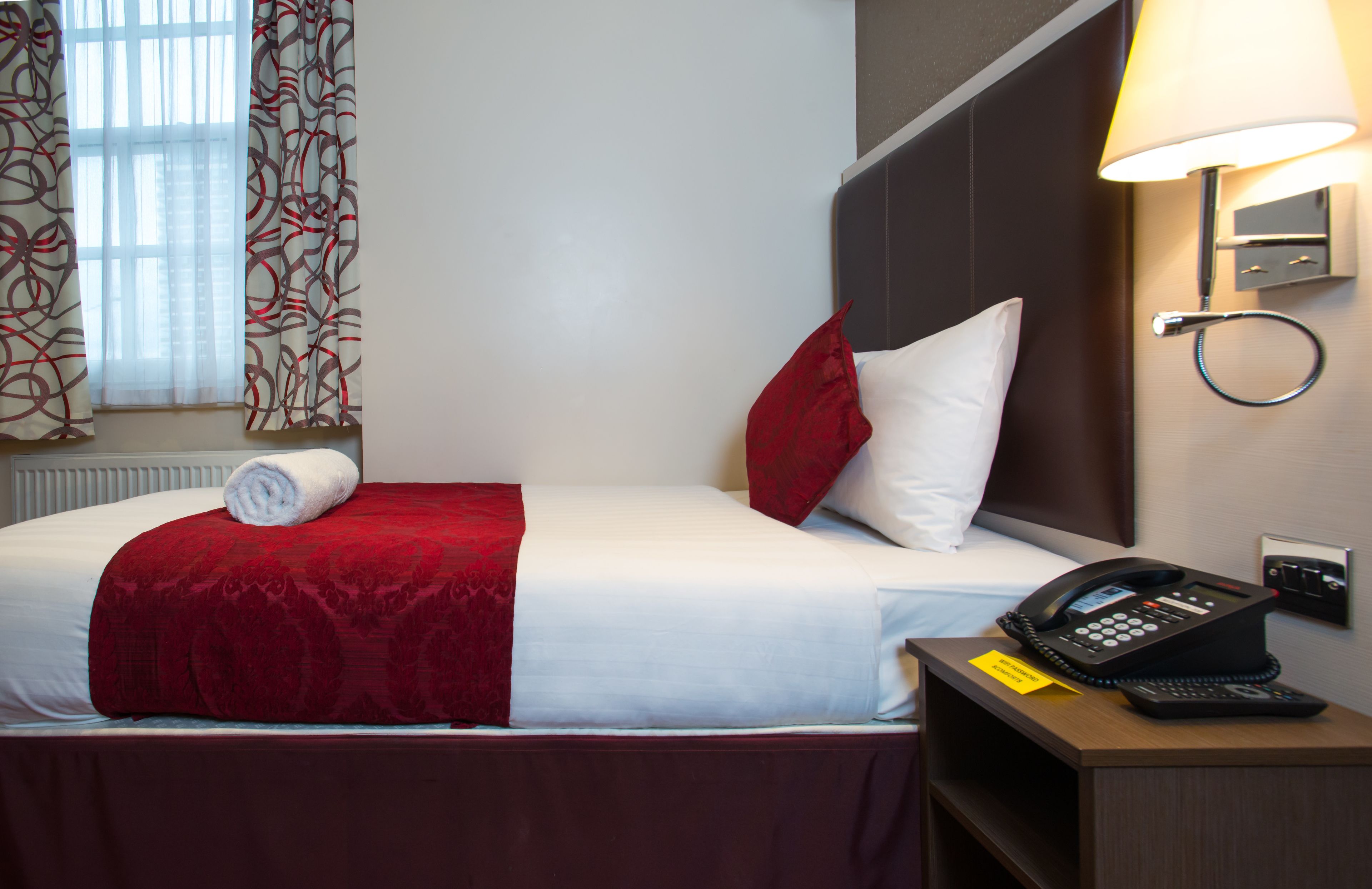 Quarto de hotel com uma cama bem arrumada, telefone ao lado da cama e mesa de cabeceira com luminária.