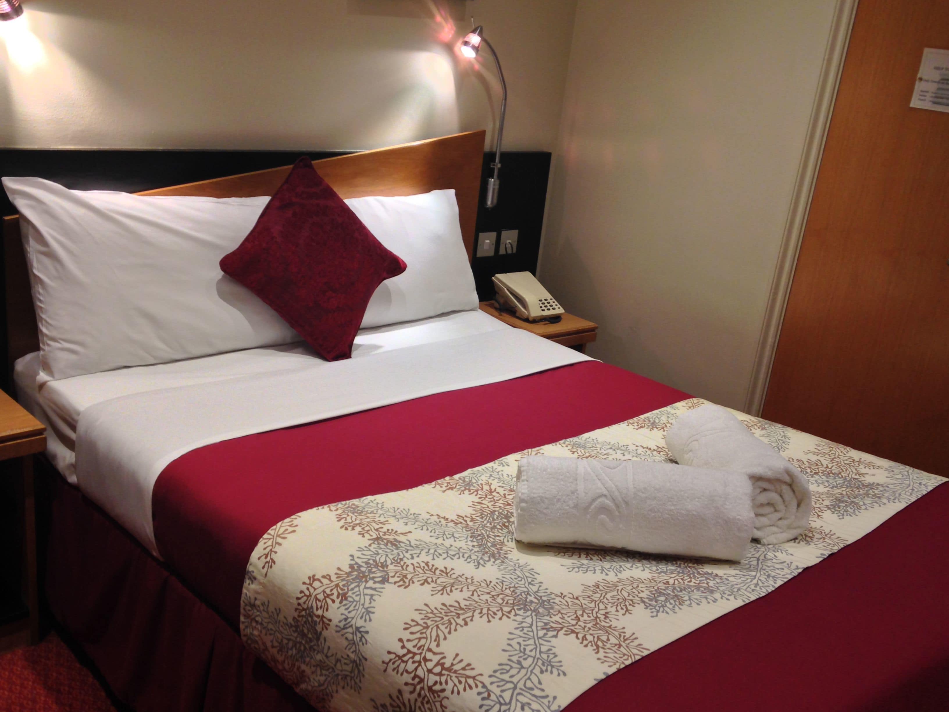 Ein Hotelzimmer mit einem ordentlich bezogenem Bett, einer roten und weißen Tagesdecke, einem roten Kopfkissen und einem weißen Telefon auf einem Nachttisch. Eine Wandlampe vervollständigt das Bild.