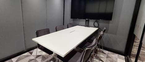Un salón de conferencias con una gran mesa blanca, seis sillas y un televisor de pantalla plana montado en la pared.