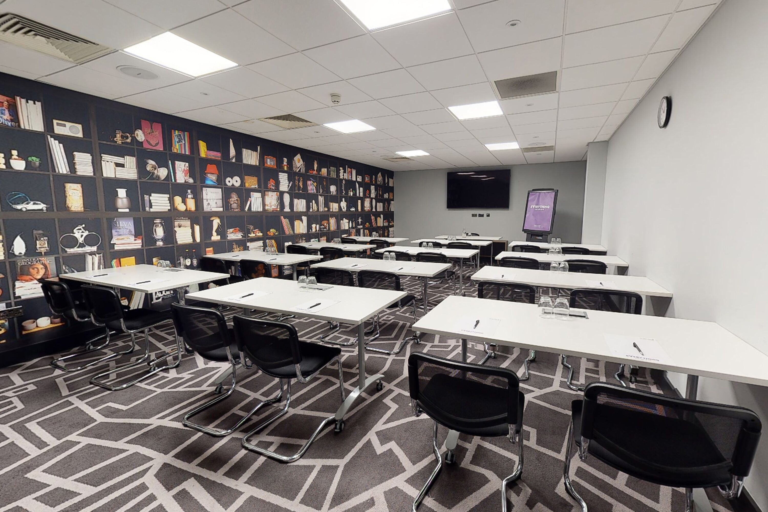 A conference room with multiple tables and chairs, a large screen, and a wall decorated with various items.