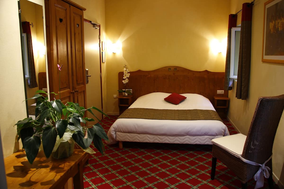 A hotel room with a wooden headboard, a single bed with a red pillow, a nightstand, a potted plant, and a wooden chair.