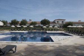 Une piscine entourée de chaises longues et de parasols, elle-même bordée d’un espace paysager et avec des bâtiments en arrière-plan.