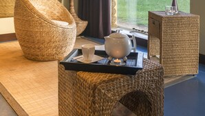 Een gezellige kamer met rieten meubels, een dienblad met een theepot, kopje en glas, en uitzicht op een gebouw en gazon.