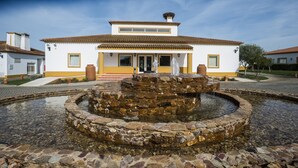 Een wit gebouw met een stenen fontein ervoor, een heldere lucht en een verzorgde binnentuin.
