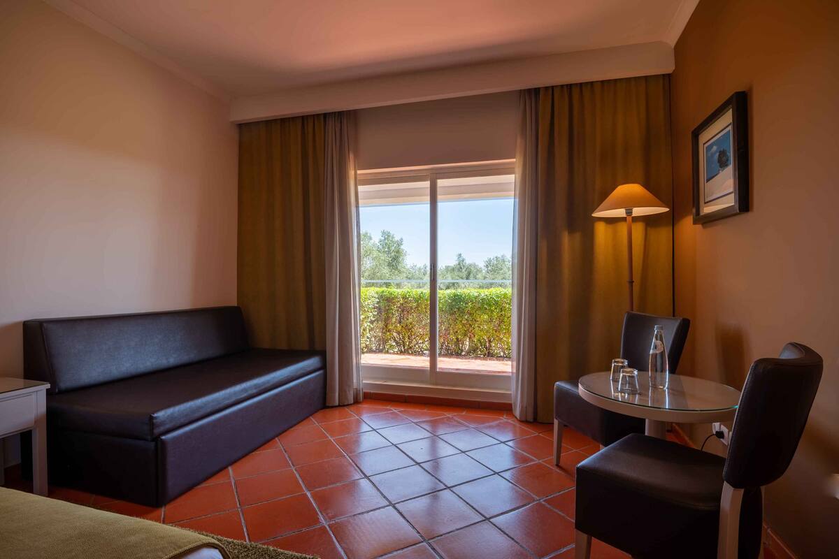 Habitación de hotel con sofá, una mesa con silla, lámpara y vistas a través de una puerta de cristal.