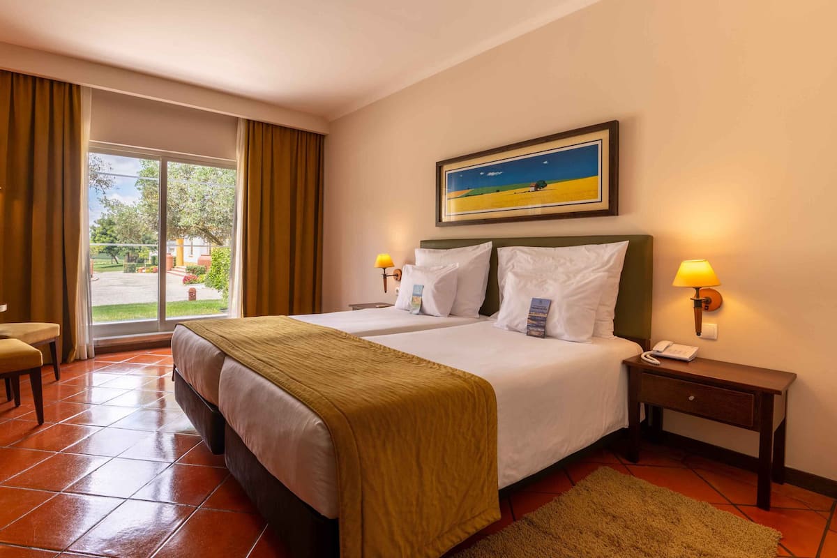 Habitación de hotel con cama, dos mesitas de noche, una silla, ventana con cortinas y un cuadro en la pared.