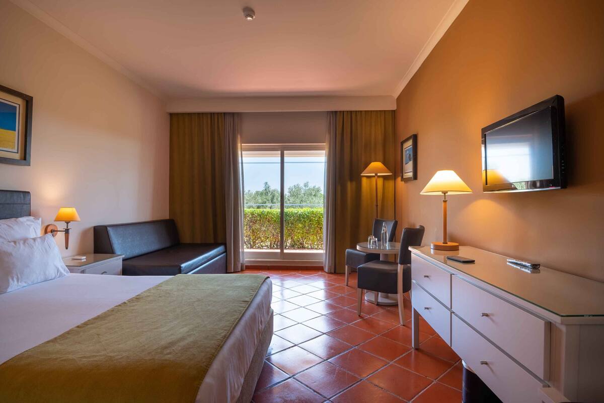 Habitación de hotel con cama, escritorio, silla, televisor y vista a la vegetación por la ventana.
