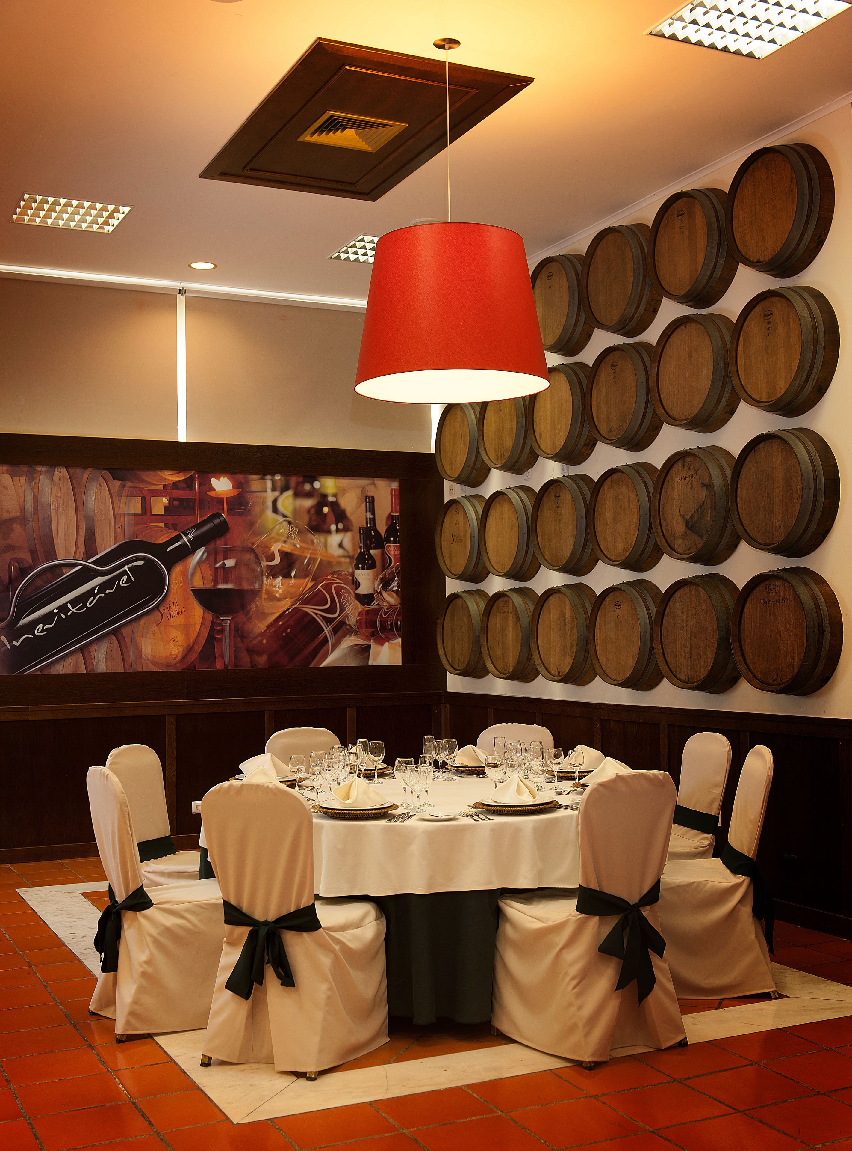 Un comedor con una mesa redonda dispuesta para seis comensales, rodeada de sillas con cintas negras, una lámpara colgante roja y una pared de barriles de madera.
