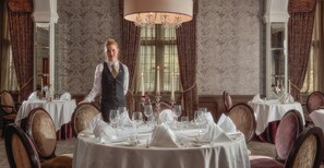 A formal dining room with multiple round tables set for a meal, a chandelier, and a waiter in uniform. - Bovey Castle (Newton Abbot)