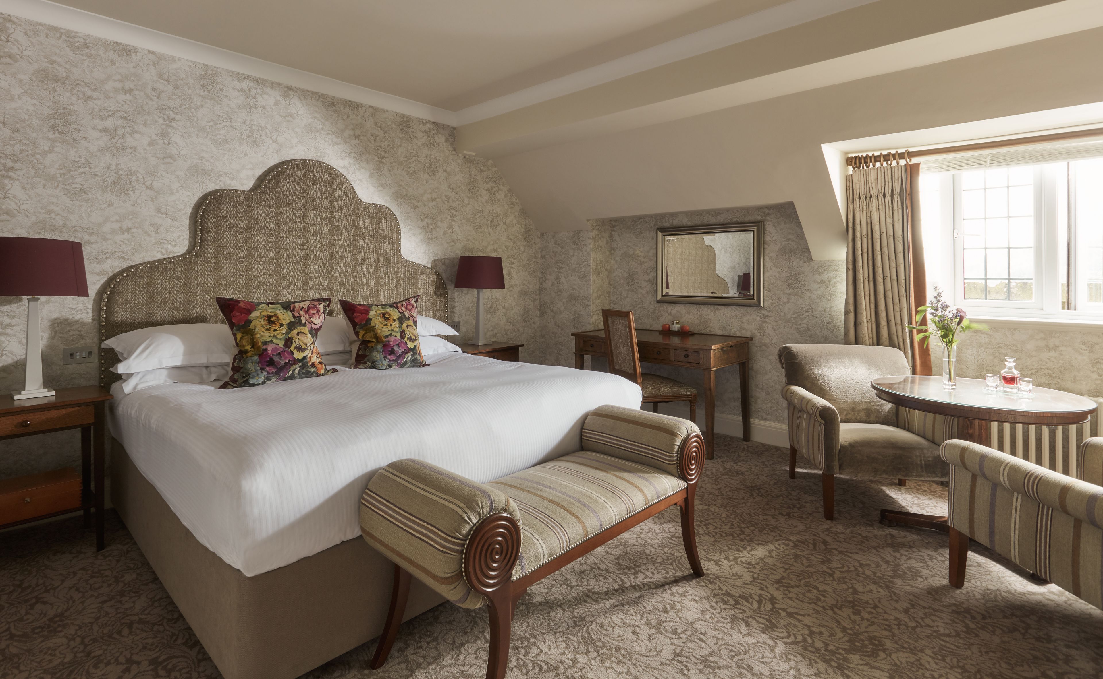 A hotel room with a large bed, a wooden chair, a small table, a desk, and a window with curtains.