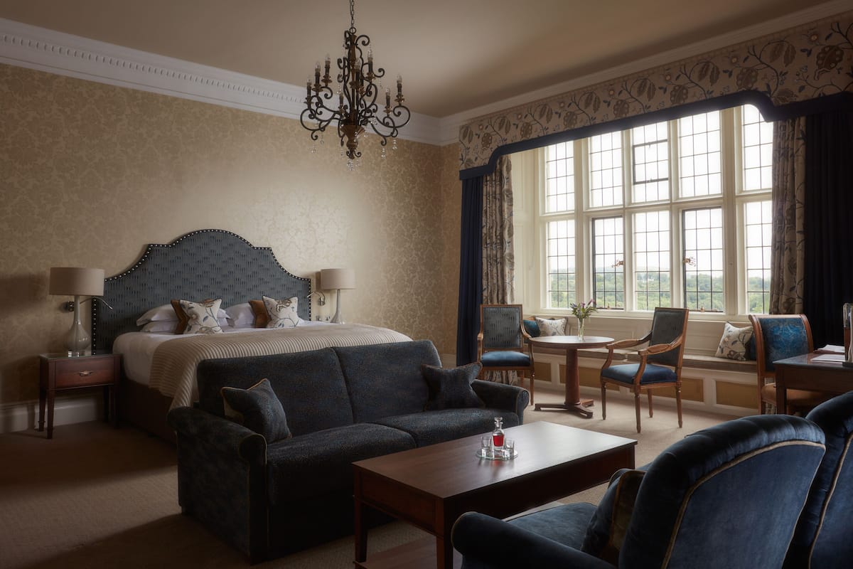 A hotel room with a large bed, a dark blue sofa, a wooden coffee table, and a view of a garden through a large window.