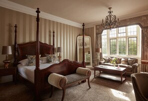 A four-poster bed with a wooden headboard, a sitting bench, a coffee table, and a sofa in a well-lit room with a large window and a chandelier. - Bovey Castle (Newton Abbot)
