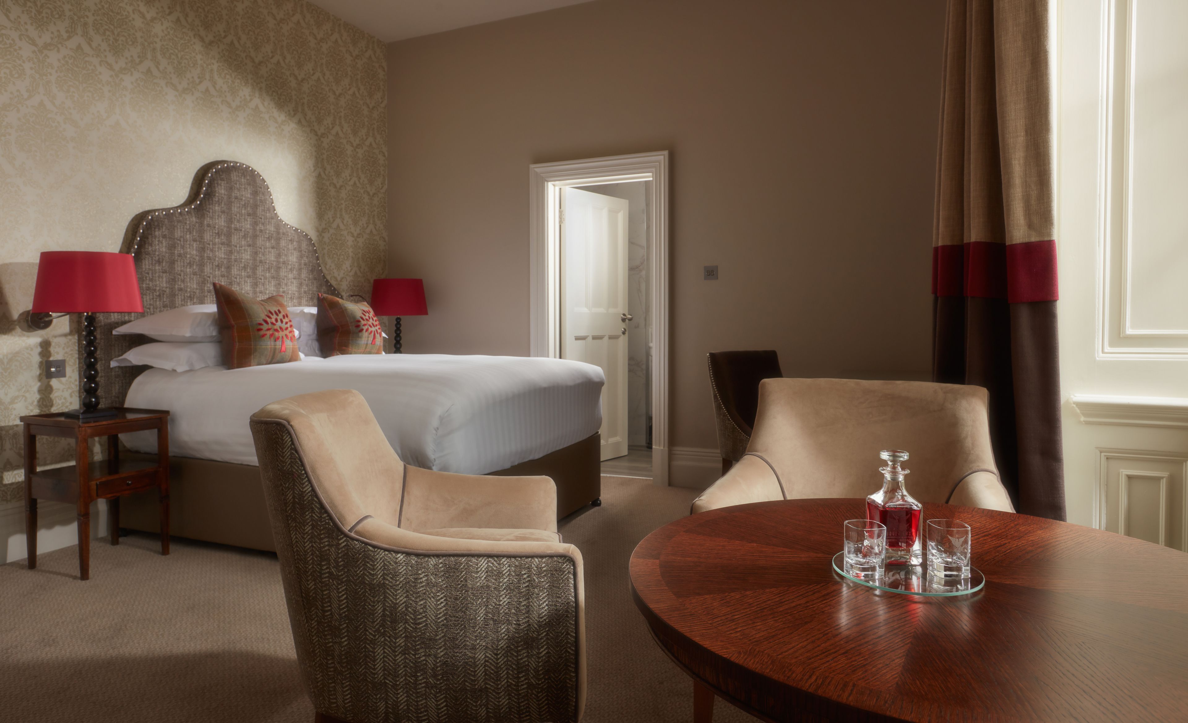 A hotel room with a bed, two chairs, a table with a bottle and glasses, and a door leading to another room.
