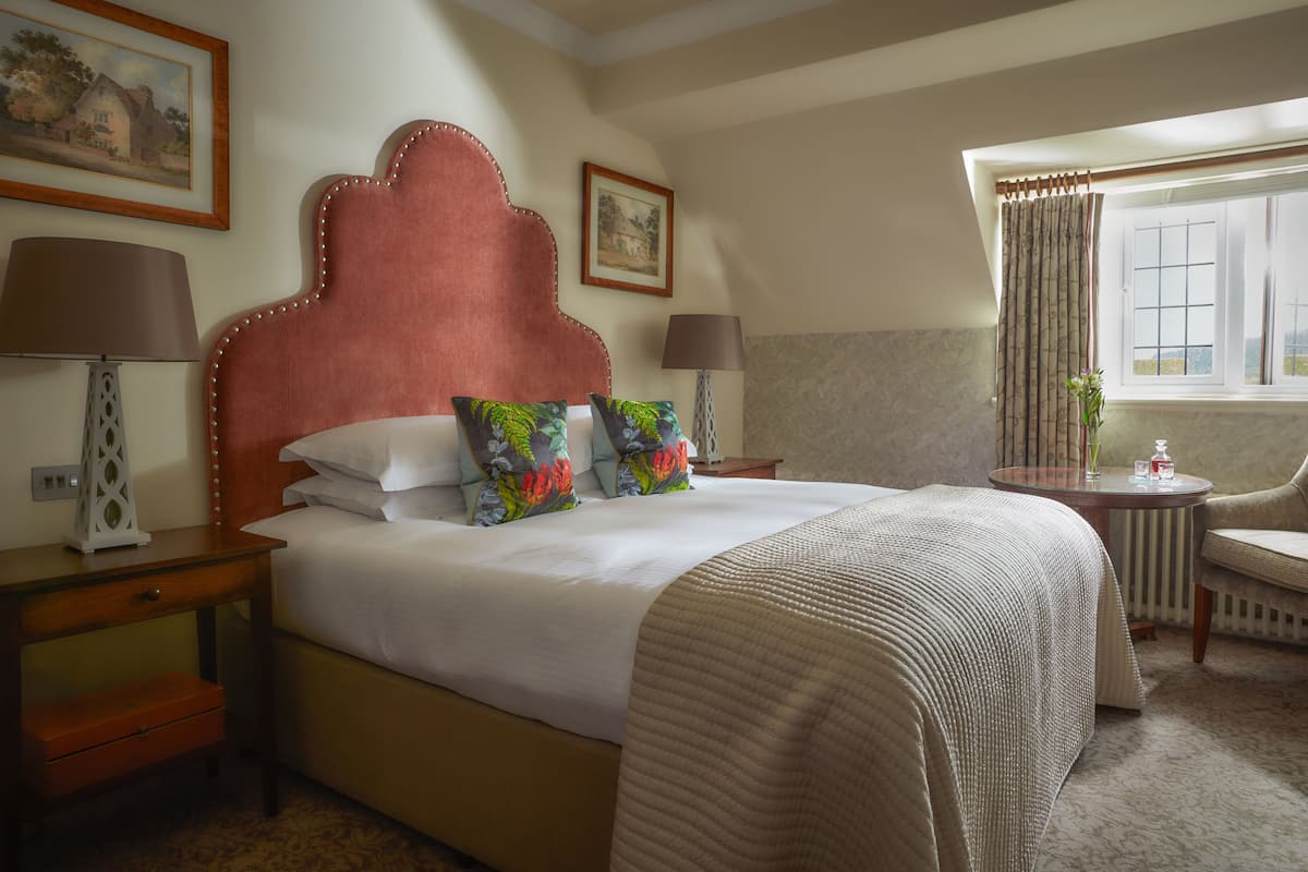 A hotel room with a large bed, two bedside tables, a chair, a window with curtains, and a painting on the wall.