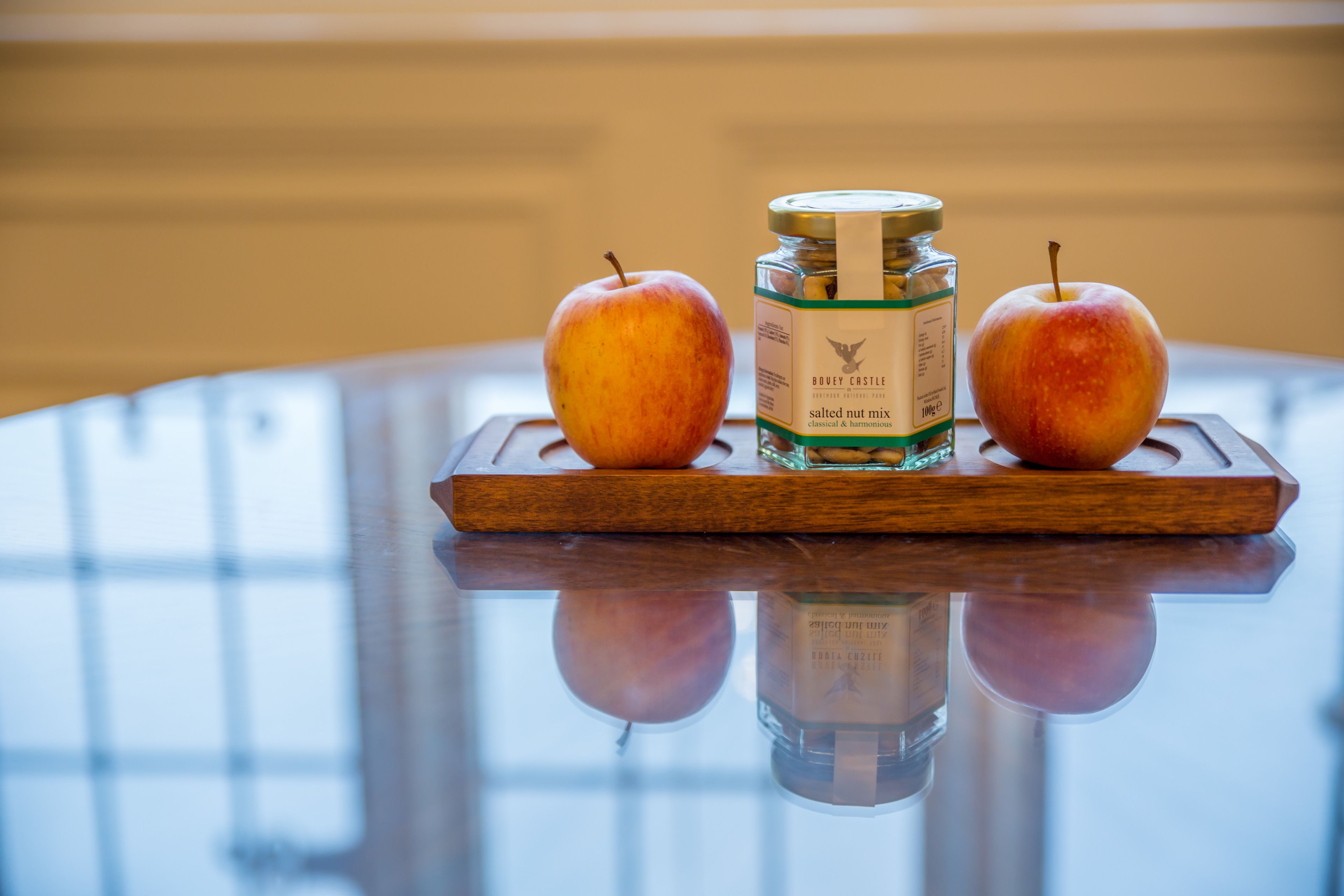 Two apples and a bottle of Boodle Castle salted nut mix on a wooden tray.