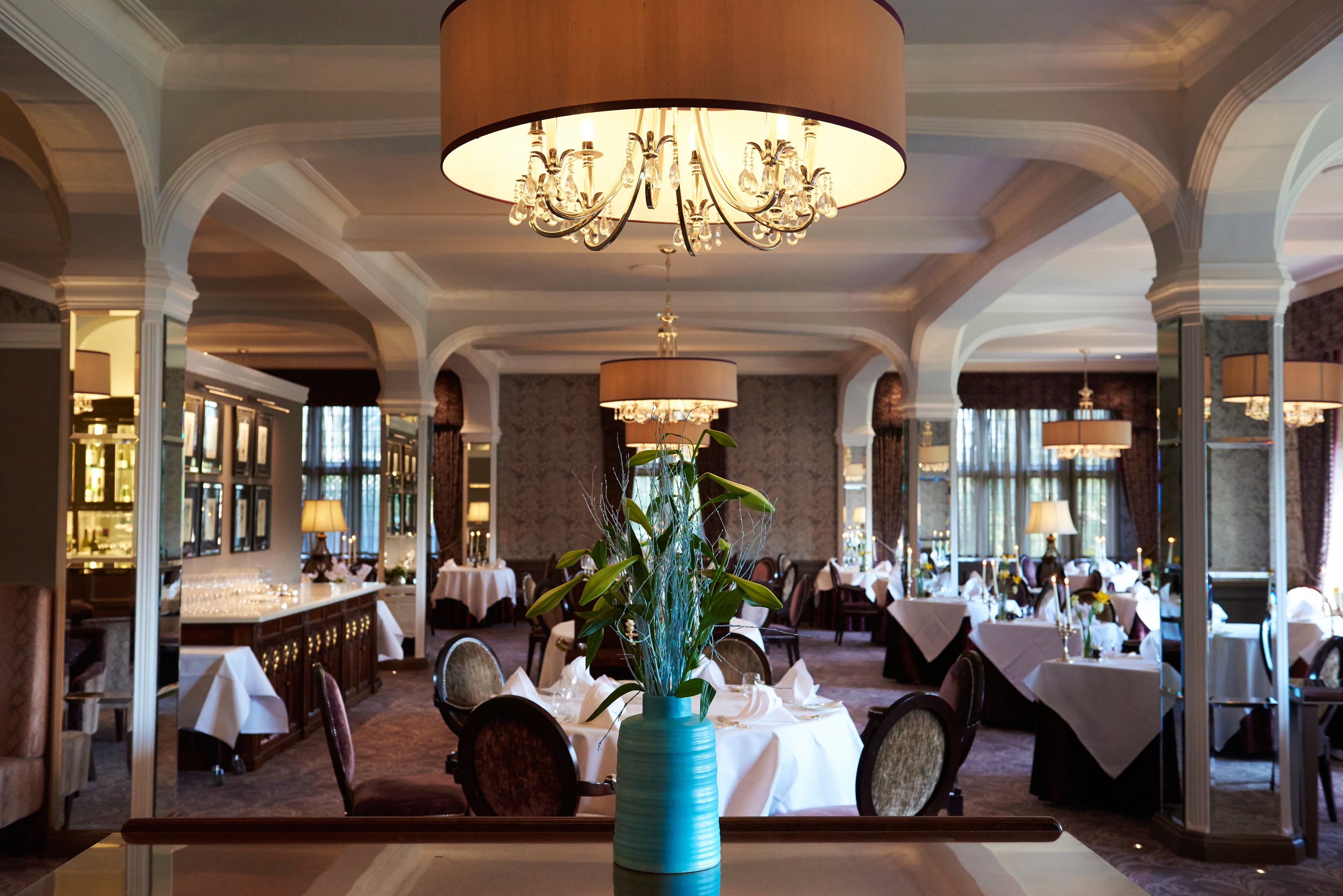 A formal dining room with tables set for service, a large chandelier, and a blue vase centerpiece.